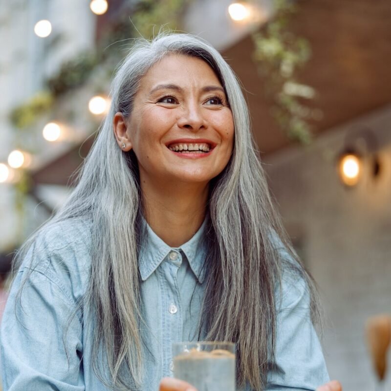 Cheerful mature Asian woman with glass of water and phone sits at small table outdoors